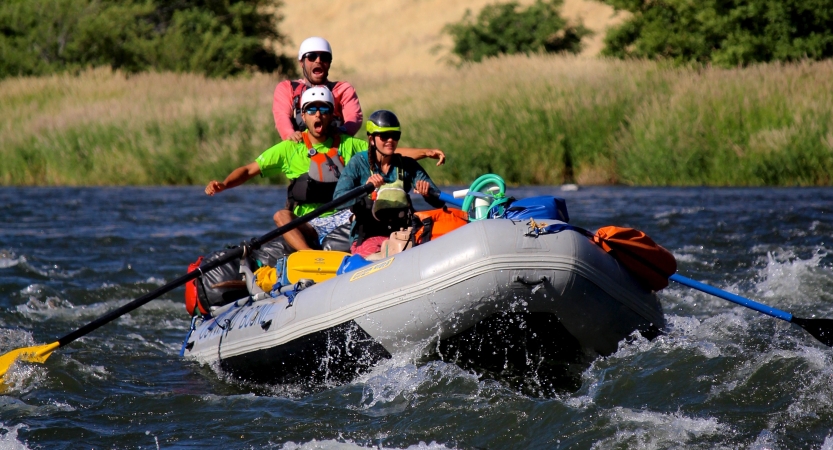 Three people wearing safety gear paddle a raft through whitewater.